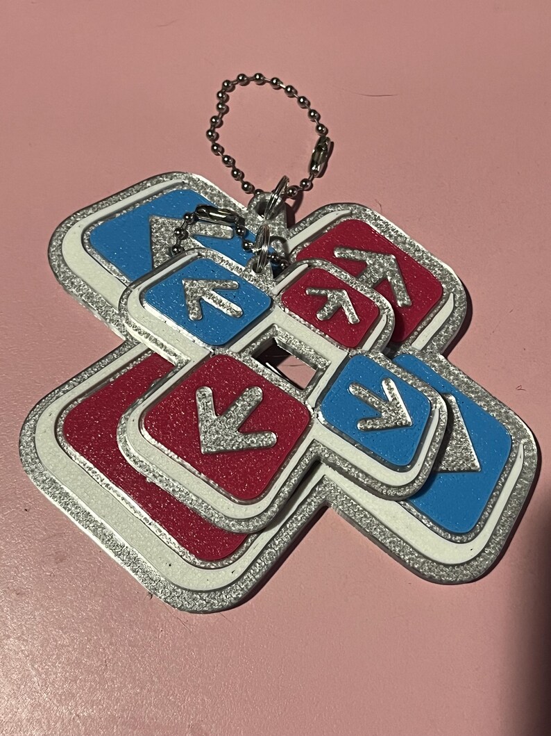 May include: A keychain featuring a layered design of red and blue squares with silver arrows and a silver border. The keychain has a silver chain for attachment. The background is a solid pink color.