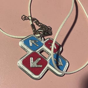 May include: A silver-colored pendant necklace with a white cord. The pendant is a cross-shaped design with red and blue squares, each featuring a silver arrow. The necklace is likely a fashion accessory.