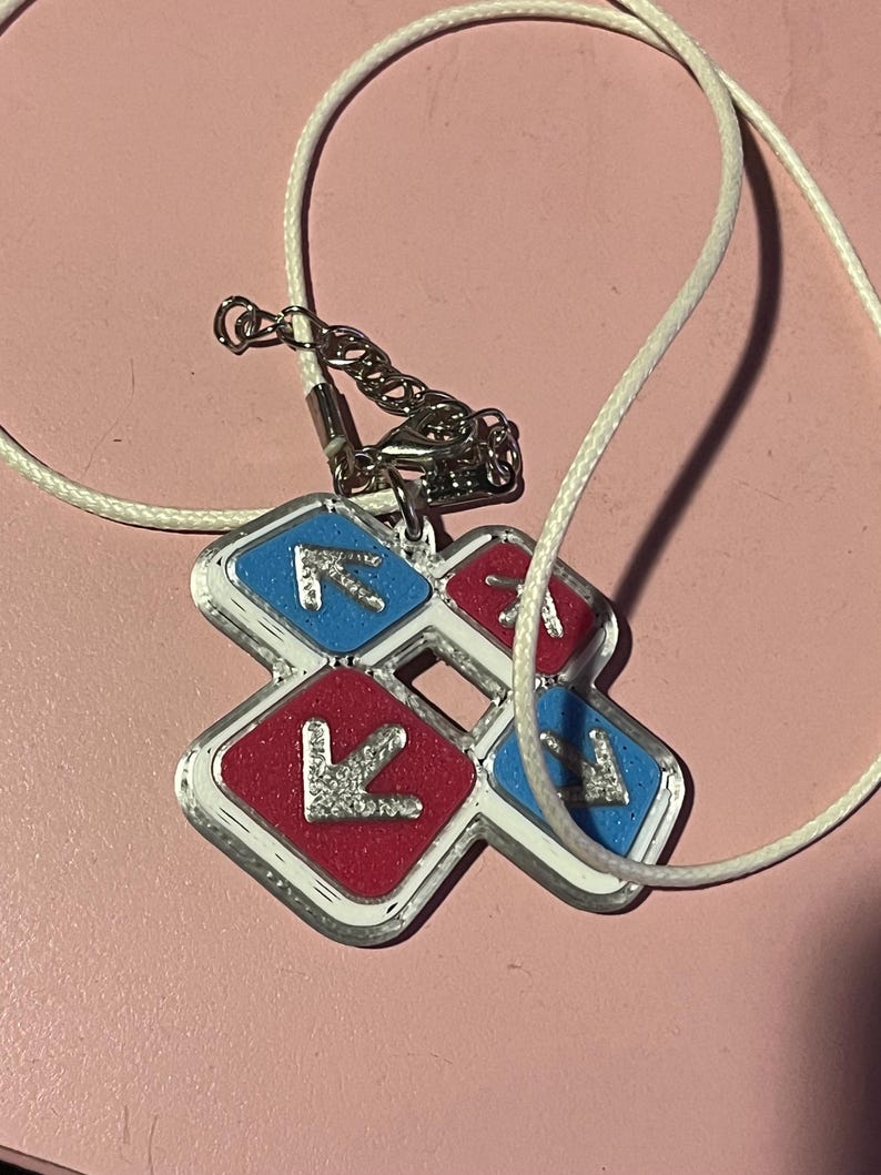 May include: A silver-colored pendant necklace with a white cord. The pendant is a cross-shaped design with red and blue squares, each featuring a silver arrow. The necklace is likely a fashion accessory.