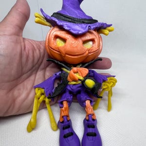 Puede incluir: Una figura de Halloween impresa en 3D de una calabaza con un sombrero morado y brazos y piernas amarillos. La calabaza tiene una cara tallada con pintura naranja y amarilla.