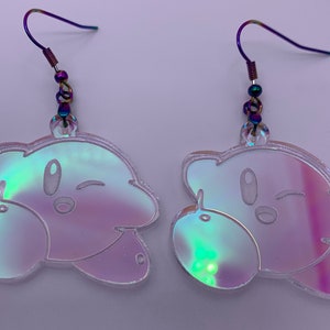 May include: A pair of iridescent pink and blue earrings featuring a cartoon character with a winking face. The earrings are made of acrylic and have a silver hook.
