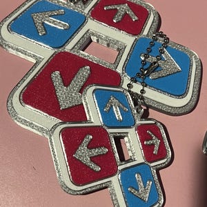 May include: A cluster of keychains featuring a dance pad design. Each pad has a red or blue background with a silver arrow. The edges are outlined in silver glitter and connected by a silver chain.