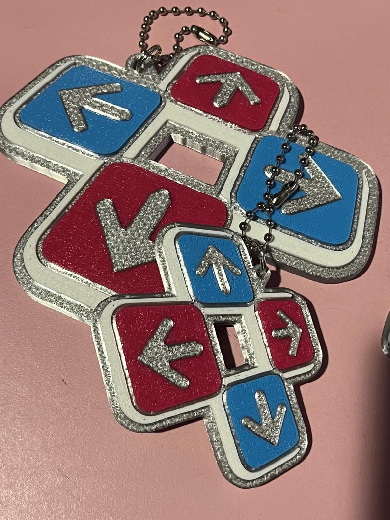 May include: A cluster of keychains featuring a dance pad design. Each pad has a red or blue background with a silver arrow. The edges are outlined in silver glitter and connected by a silver chain.