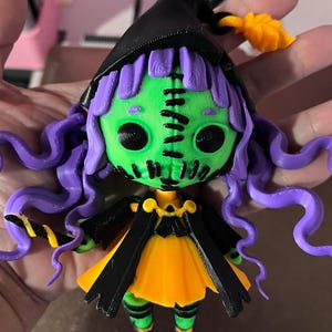May include: A green and purple witch doll with black and yellow clothing and a black witch hat. The doll has a stitched face and large black eyes.