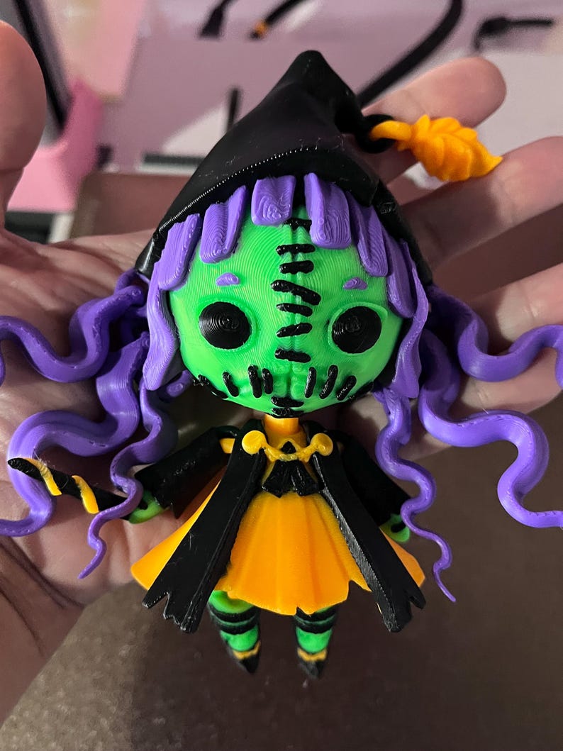 May include: A green and purple witch doll with black and yellow clothing and a black witch hat. The doll has a stitched face and large black eyes.