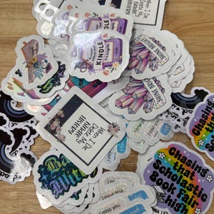 May include: A collection of stickers with various designs and text. Some stickers feature crystals, books, and the text "Shine Bright." Other stickers have phrases like "When I Die Delete My Kindle History" and "Chasing that Scholastic Book Fair high."