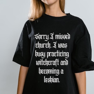 May include: A black short-sleeved t-shirt featuring white text in a gothic-style font that reads: Sorry I missed church. I was busy practicing witchcraft and becoming a lesbian. The shirt has a crew neckline and a relaxed, loose-fitting silhouette.