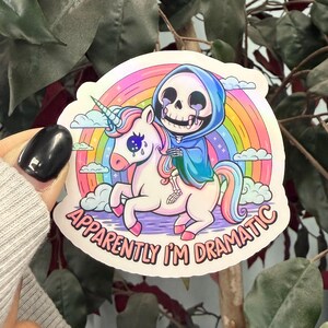 Könnte beinhalten: Ein Aufkleber mit einem Cartoon-Sensenmann, der auf einem Einhorn über einem Regenbogen reitet. Der Sensenmann trägt einen blauen Kapuzenmantel und weint. Das Einhorn hat eine rosa Mähne und einen rosa Schweif. Der Text auf dem Aufkleber lautet "Apparently I'm Dramatic."