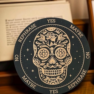 May include: A round, dark teal wooden board with a detailed, engraved sugar skull design. The skull is surrounded by the words "YES", "MAYBE", "NO", and "REPHRASE". Stars and dots complete the design.