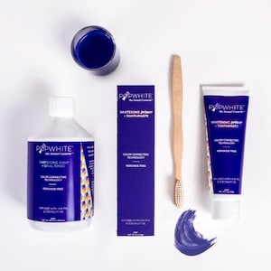 May include: A flat lay of dental hygiene products, including a bottle of oral rinse, a tube of toothpaste, a wooden toothbrush, and a small glass of blue liquid. The products are white and blue, with the brand name "Popwhite" visible.