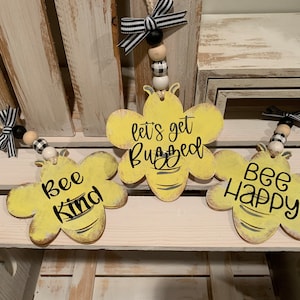 May include: Three yellow wooden bee-shaped ornaments with black lettering. The ornaments are hanging from twine with black and white checkered ribbon and wooden beads. The text on the ornaments reads "Bee Kind", "Let's Get Buzzed", and "Bee Happy".