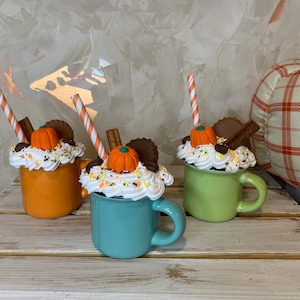 May include: Three ceramic mugs decorated with whipped cream, chocolate, sprinkles, and a miniature pumpkin. Each mug has a straw and is a different color: orange, teal, and green.