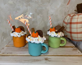 May include: Three ceramic mugs decorated with whipped cream, chocolate, sprinkles, and a miniature pumpkin. Each mug has a straw and is a different color: orange, teal, and green.
