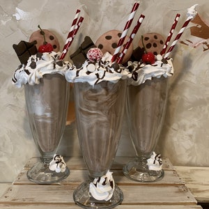 May include: Three chocolate milkshakes with whipped cream, chocolate syrup, chocolate chips, cherries, and cookies. Each milkshake has a red and white striped straw.