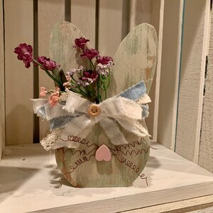 May include: A wooden bunny shaped planter with a distressed green paint finish. The planter is decorated with a pink heart, a button, and a bow made of white and blue fabric. The planter is filled with pink and purple flowers.