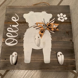 May include: A wooden dog leash holder with two white hooks. The holder features a white silhouette of a dog with a bow and the name "Ollie" written in white cursive.