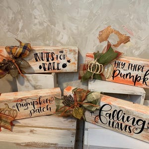 May include: Wooden fall-themed signs with phrases like "Nuts for Fall" and "Hey There Pumpkin." Each sign is decorated with a bow and a pumpkin. The signs are painted in orange and white, with black lettering.
