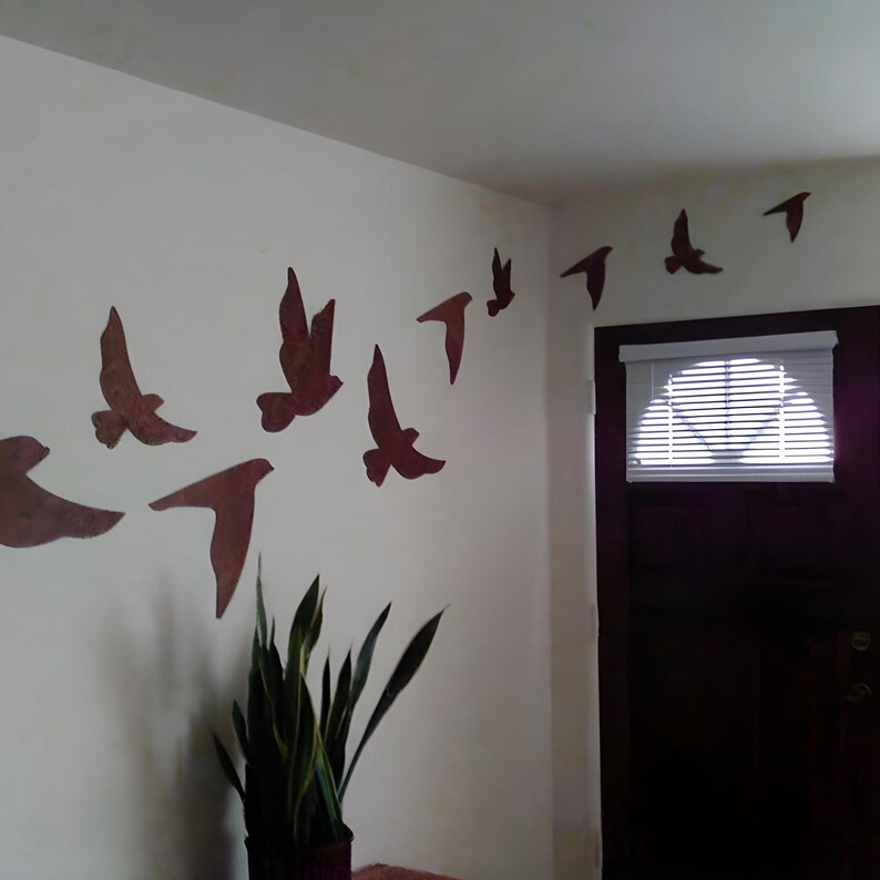 May include: A white wall with a collection of brown metal bird silhouettes in flight. The birds are arranged in a diagonal line, starting from the top right corner of the wall and extending down to the left. A potted plant sits on the floor below the birds.