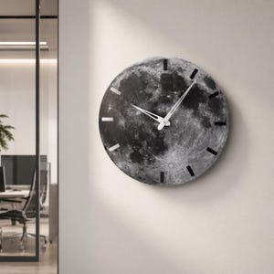 May include: A round wall clock featuring a detailed image of the moon's surface. The clock face is primarily black and white, with white clock hands and black and white hour markers. The clock is mounted on a light-colored wall.