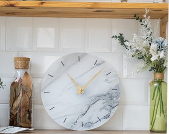 May include: A white marble clock with gold hands and black markings. The clock is round and has a white background with gray marble veins.