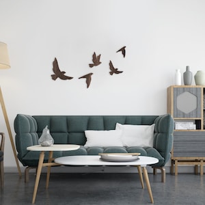 May include: A teal sofa with white pillows sits in a living room with a wooden floor. A wooden floor lamp with a white lampshade is in the corner. A wooden coffee table with a white top sits in front of the sofa. A wooden cabinet with a gray door and shelves is in the background. The wall is white with a flock of brown birds in flight.