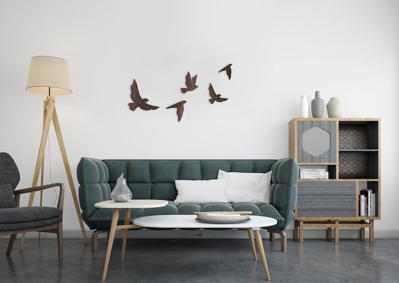 May include: A teal sofa with white pillows sits in a living room with a wooden floor. A wooden floor lamp with a white lampshade is in the corner. A wooden coffee table with a white top sits in front of the sofa. A wooden cabinet with a gray door and shelves is in the background. The wall is white with a flock of brown birds in flight.