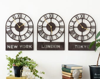 Puede incluir: Tres relojes de pared decorativos con marcos de metal negro y esferas con números romanos. Los relojes están etiquetados como "Nueva York", "Londres" y "Tokio".