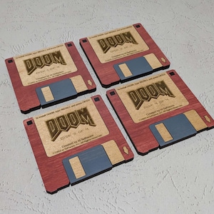 May include: Four wooden coasters shaped like floppy disks with the text "DOOM" and "DISK # OF 4" printed on them. The coasters are red with a wood grain texture and have a blue and brown floppy disk design.