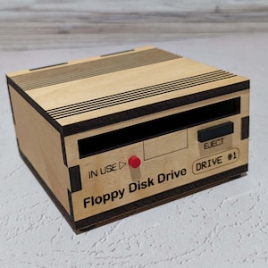 May include: A wooden Floppy Disk Drive box with laser-etched details. The box features the words "Floppy Disk Drive" and "Drive #1", along with an "EJECT" button and an "IN USE" indicator light. The box is made of light-colored wood with dark brown accents.