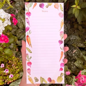 Peut inclure: Un bloc-notes ligné avec une bordure de légumes à l'aquarelle. La bordure présente des carottes, des radis, des pois et d'autres légumes du jardin. Le mot "Notes" est imprimé en haut. Le bloc-notes est tenu devant un fond de fleurs et de verdure.