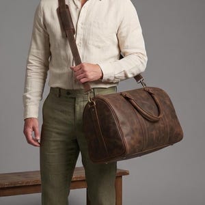 May include: A brown leather duffel bag featuring a smooth, distressed texture, two top handles, and a long adjustable shoulder strap with a padded section. The bag is constructed with reinforced stitching and metal hardware clips on the sides.