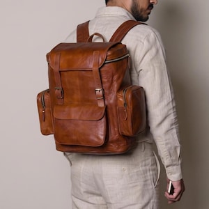 Puede incluir: Una mochila de cuero color canela con un compartimento principal grande con solapa, dos correas verticales con hebillas, un bolsillo frontal central y dos bolsillos laterales con cierres de cremallera. Incluye correas ajustables.