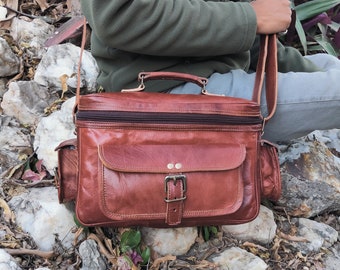 May include: A brown leather satchel with a top handle, shoulder strap, and multiple pockets. The bag features a buckle closure on the front flap and is designed for carrying items. The satchel is shown outdoors, resting on a person's lap.