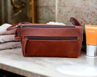 May include: A brown leather travel toiletry bag with two zippered compartments and a small zippered pocket on the front. The bag is sitting on a white marble countertop.
