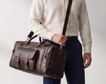 Pode incluir: Uma mala de viagem em couro castanho escuro com um bolso frontal grande com duas correias de fivela, duas pegas superiores e uma alça de ombro destacável com ferragens metálicas. A mala tem uma forma retangular com costuras visíveis.