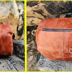 May include: A brown leather duffel bag with two top handles and a zipper closure. The bag is sitting on a rocky surface.