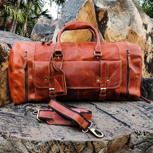 May include: A brown leather duffel bag with a long strap and a smaller pocket on the front. The bag is sitting on a large rock.