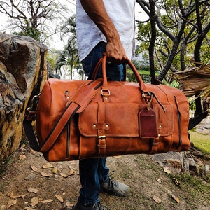 May include: A brown leather duffel bag with a strap and a luggage tag. The bag is being held by a person wearing blue jeans.