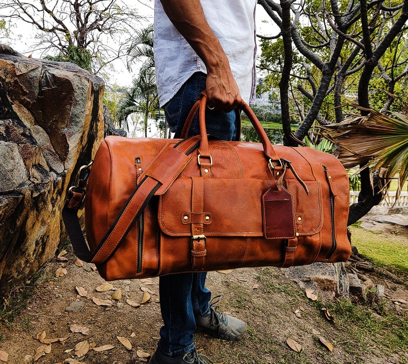 May include: A brown leather duffel bag with a strap and a luggage tag. The bag is being held by a person wearing blue jeans.