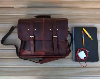 May include: A brown leather messenger bag with a long adjustable strap, two buckles, and a flap closure. The bag is sitting on a wooden surface next to a laptop, a charger, and a few pens.