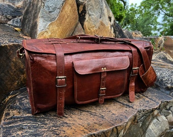 Puede incluir: Una bolsa de viaje de cuero marrón con múltiples hebillas y un gran bolsillo frontal. La bolsa está sentada sobre una superficie rocosa.