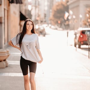 May include: A light gray t-shirt with the text "Keep your spirits when you're alive" and a floral skull design. The person is wearing black biker shorts and flip-flops. The background is a city street.