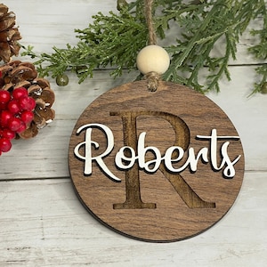 May include: A round wooden Christmas ornament with a white background and the name 'Roberts' written in a cursive font. The letter 'R' is cut out of the wood and the rest of the name is layered on top. The ornament is hanging from a twine string with a wooden bead.