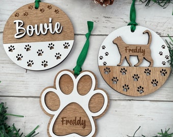 Puede incluir: Tres adornos navideños de madera con diseños de huellas de patas. Un adorno es un círculo con el nombre "Bowie" y huellas de patas. Otro adorno es un círculo con una silueta de gato y el nombre "Freddy". El tercer adorno es una huella de pata con el nombre "Freddy".