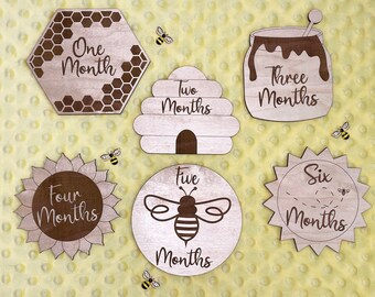 May include: Six wooden baby milestone cards with a bee theme. The cards feature a honeycomb, beehive, honey jar, sunflower, bee, and sun design. Each card has a number and the word "months" written on it.