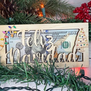 May include: A wooden Christmas card with the words "Feliz Navidad" and a cutout for a banknote. The card also features a small Christmas tree with colourful lights and a string of lights.