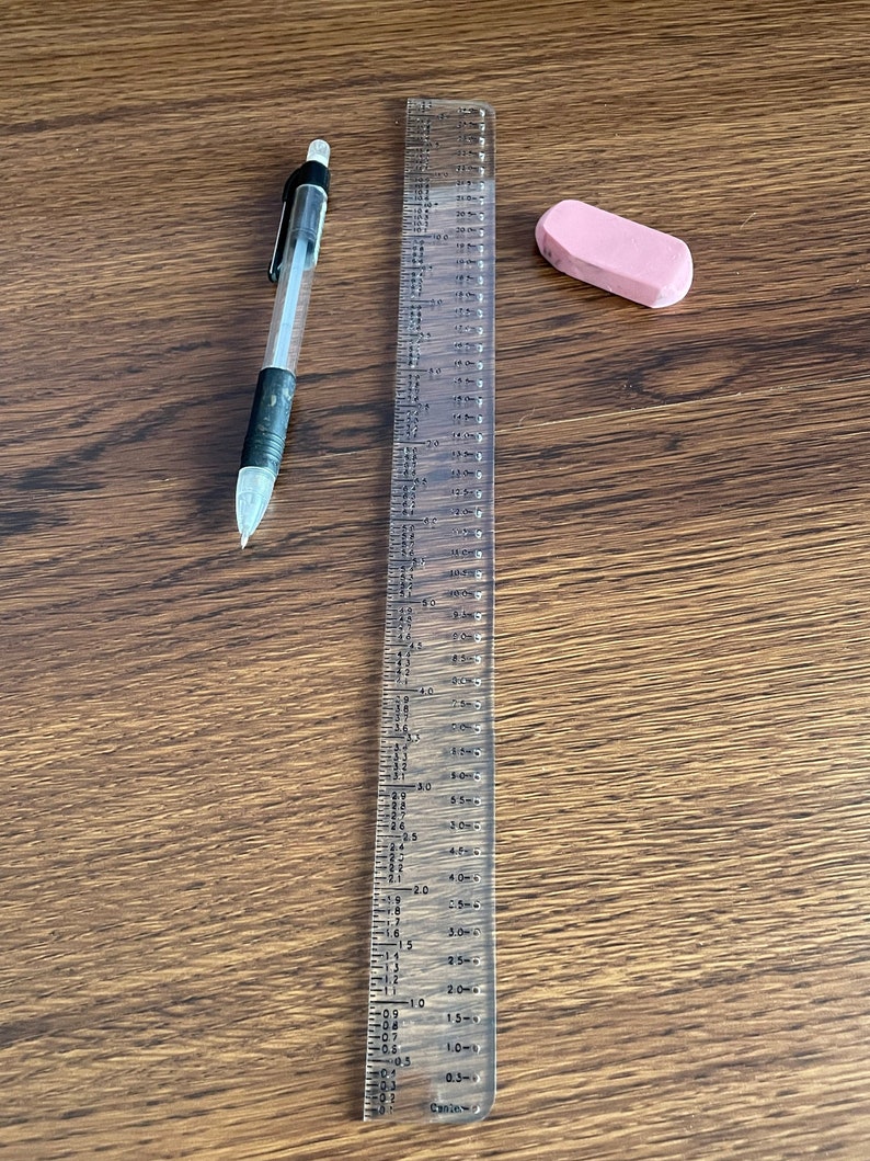 K&ouml;nnte beinhalten: Ein durchsichtiges Lineal aus Kunststoff mit schwarzen Markierungen, ein rosa Radiergummi und ein schwarzer und transparenter Bleistift auf einer braunen Holzoberfl&auml;che.