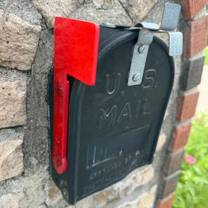 May include: Black U.S. Mail mailbox with a bright red flag, mounted on a stone wall. The mailbox has the words "U.S. MAIL" embossed on the front. The flag is in the upright position.