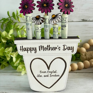 May include: Wooden planter with a heart-shaped cutout and the text "Happy Mother's Day" and "From Crystal. Alex, and Brecken". The planter has three pink flowers with white daisy flowers in the center. The flowers are attached to rolled up dollar bills.