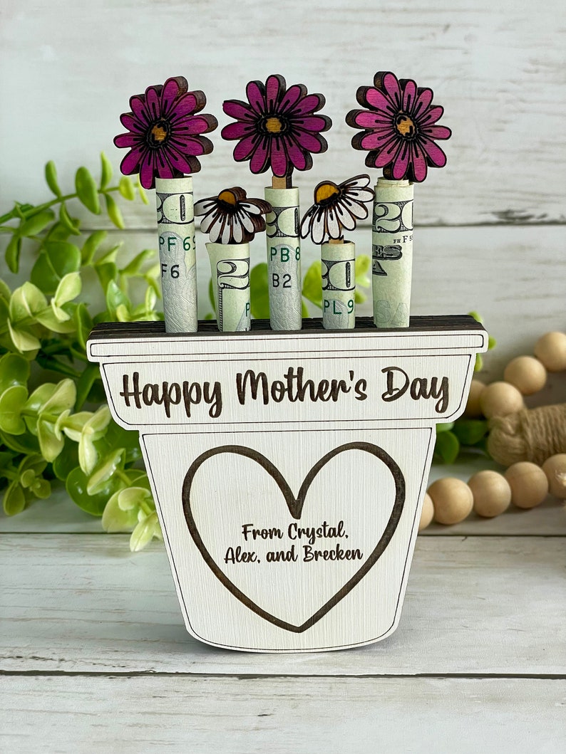 May include: Wooden planter with a heart-shaped cutout and the text "Happy Mother's Day" and "From Crystal. Alex, and Brecken". The planter has three pink flowers with white daisy flowers in the center. The flowers are attached to rolled up dollar bills.